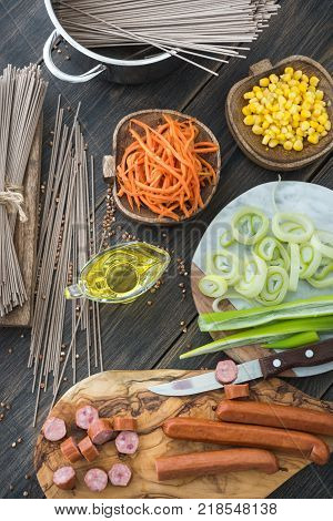 Slicing sousages for soba noodles cooking with leek, corn and korean carrot on a dark wooden rustic background top view
