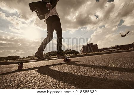 People youth urban lifestyle extreme sports and activity concept. Outdoor action shot of stylish young male in sneakers performing tricks using long board casting shadow of pavement. Warm filter