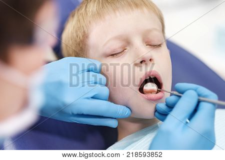 Male dentist examines the teeth of the patient cheerful child with blond hair. Boy smiling in dentist's chair with mouth wide open.