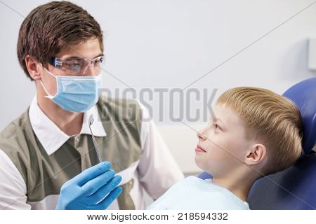 Male dentist examines the teeth of the patient cheerful child with blond hair. Boy smiling in dentist's chair with mouth wide open.