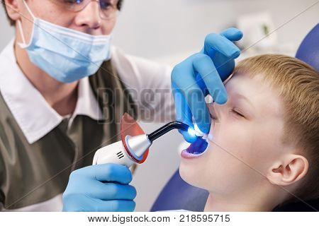 Male dentist examines the teeth of the patient cheerful child with blond hair. Boy smiling in dentist's chair with mouth wide open.