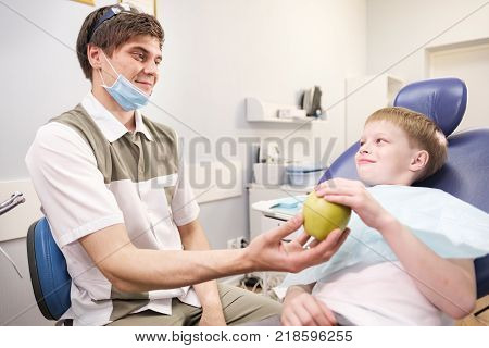 Doctor male dentist giving green apple to patient boy in dental chair