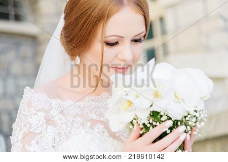 Smiling Young bride in white dress is sniffing her wedding bouquet which is carefully held by hands. The concept of florists at wedding events