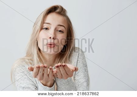 Blonde appealing female blows kiss at camera, demonstrates love to boyfriend or says goodbye, posing against gray studio background. Attractive young woman with dark eyes shows sympathy to someone
