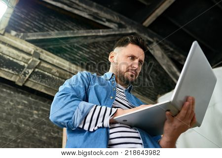 Low angle portrait of handsome mature  man using laptop while standing in barn-like workshop, copy space