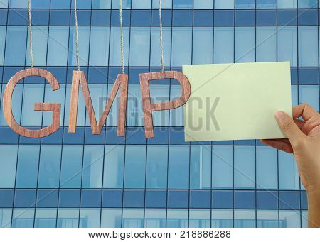 Wooden GMP text Stands for Good Manufacturing Practice hanging on rope and have hand of a businessman holding a green note on Skyscraper background.
