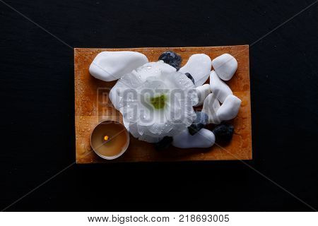 A bud of a white beautiful flower, a small burning candle and a multiform pebble, for spa treatments on a small wooden board. On a black background. View from above.