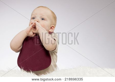 Excited toddler baby looking up and holding his palms near his mouth. Idea for greeting card. Surprised infant boy sitting on the white blanket. Horizontal studio shot. Copy space