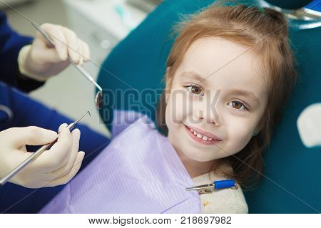 Child with cute smile sits at dentist chair with napkin on chest and doctor in sterile rubber gloves trys to start examination with special medical instruments.