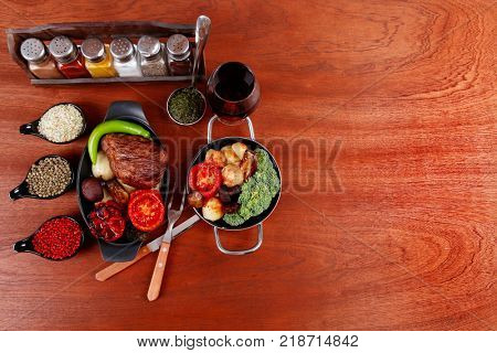 fresh roast beef meat rib eye steak with tomatoes,potatoes, hot chili pepper gern and pink peppercorn saucer, cutlery, set of spice, and red wine glass on light walnut table