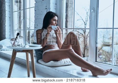 Relaxing at home. Attractive young woman looking away through the window and having morning coffee while sitting on the window sill at home