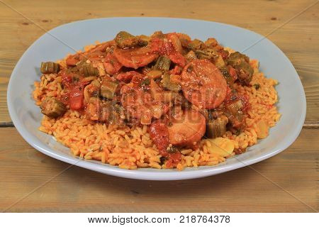 Okra cooked with tomatoes sausage ingredients and seasonings served over Mexican style cooked white long rice in gray porcelain dish over wooden kitchen table