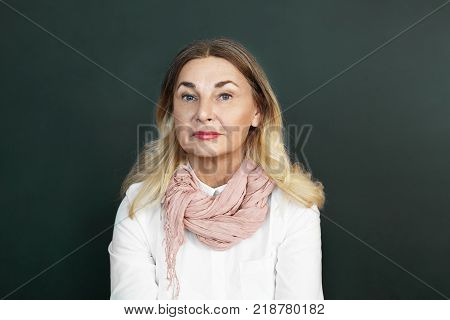 People age and lifestyle concept. Picture of beautiful Caucasian retired woman with loose blonde colored hair blue eyes and wrinkles posing in studio wearing stylish mustard scarf over white blouse