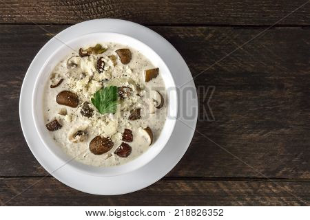 An overhead photo of a plate of a mushroom cream soup with champignons, cremini, fresh parsley, with grated cheese, on a dark rustic background texture, with a place for text