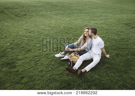 A boy and girl sitting on the green grass in the park with some food in the basket and enjoying the company of each other. He is kissing her and it's so touching