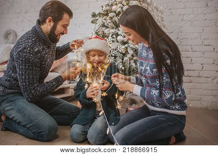 Beautiful young family enjoying their holiday time together, decorating Christmas tree, arranging the christmas lights and having fun.