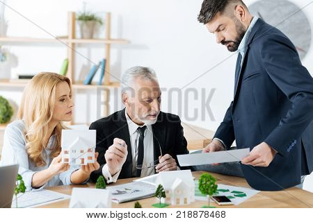 Checking details. Smart qualified real estate agent sitting on the table and looking serious while showing an important document to his reliable experienced colleagues