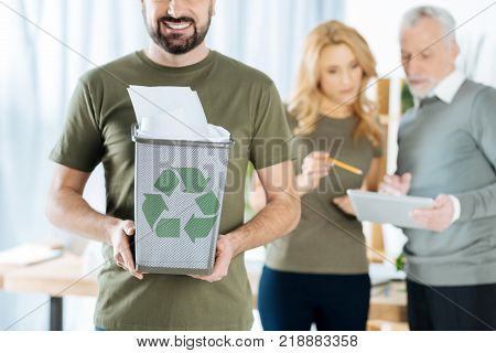 Recycling paper. Happy young man showing a nice bin full of recycled paper with a recycling symbol on it while his colleagues standing near and working