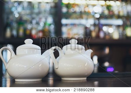 Bar counter with two white teapots, blurred alcohol bottles assortment on background. Barroom in restaurant, hotel, pub, cafe, copy space.