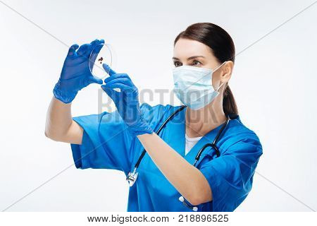 Fingerstick blood. Concentrated young female lab assistant holding  blood sample while staring  at it and dressing in mask