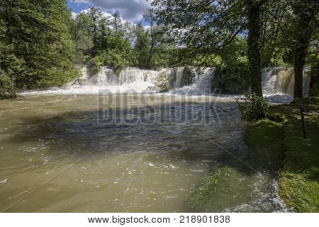 Croatian Village Of Rastoke By A River Canyon With Wooden Houses In A Green Lanscape With Trees And
