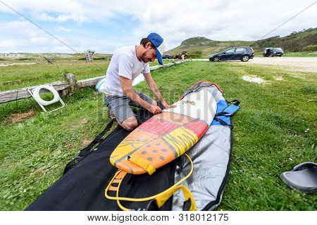 Surfer On Vacation Waxing Surf Board Outdoors On The Beach. Man Is Removing Or Applying Wax To Surfb
