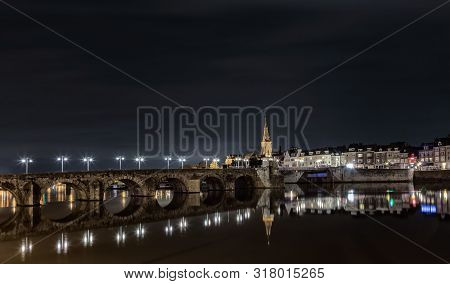 Riverside Night Scene In Maastricht, Netherlands With Old Bridge Over River Maas And Illuminated Sai