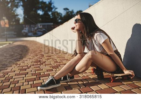 Youth, Leisure And Outdoor Activity Concept. Cheerful Dreamy Hipster Skater Girl Sit On Wooden Penny