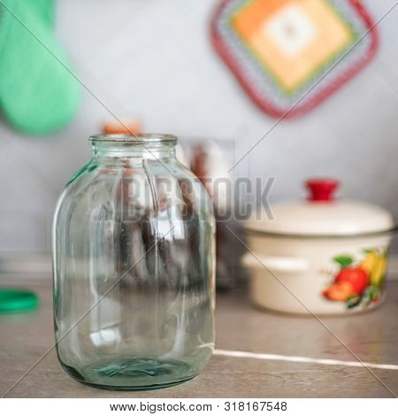 Empty Three-liter Jar Is On The Tabletop Against The Background Of The Pot And Oven Mitts.