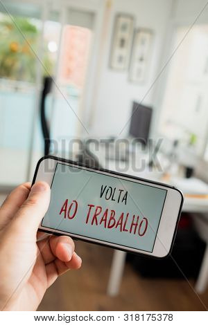 closeup of a man sitting at his office desk, and a smartphone in the foreground with the text volta ao trabalho, back to work written in portuguese, in its screen