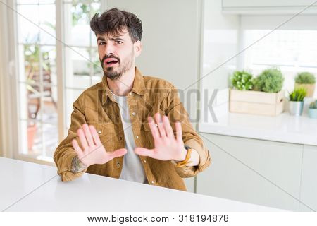 Young man wearing casual jacket sitting on white table afraid and terrified with fear expression stop gesture with hands, shouting in shock. Panic concept.
