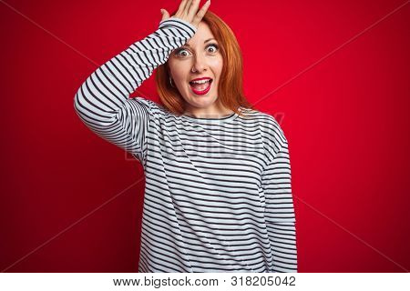 Young redhead woman wearing strapes navy shirt standing over red isolated background surprised with hand on head for mistake, remember error. Forgot, bad memory concept.