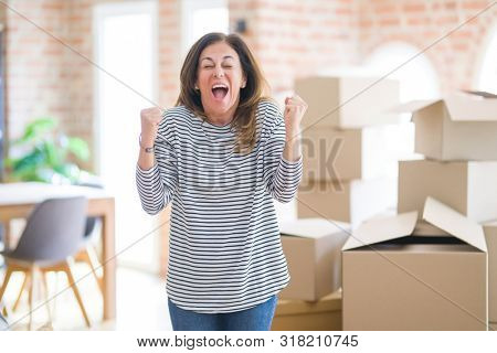 Middle age woman moving to a new house arround cardboard boxes celebrating surprised and amazed for success with arms raised and open eyes. Winner concept.