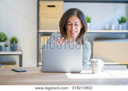 Middle age senior woman sitting at the table at home working using computer laptop disgusted expression, displeased and fearful doing disgust face because aversion reaction. With hands raised.