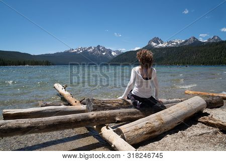 Adult Woman (30-35 Years) Sits On A Pile Of Logs On The Shoreline Of Redfish Lake In Stanley Idaho I