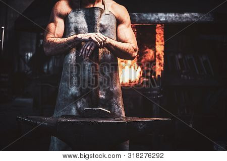 Muscular Blacksmith In Protective Apron At His Workshop With Hammer In His Hands. There Are Fire At 