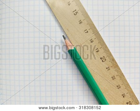 Green Pencil And Wooden Ruler With Metric System Rests On A White Checkered Exercise Book Without No