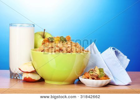 leckere Cornflakes in grüner Schale, Äpfel und Glas Milch auf Holztisch auf blauem Hintergrund