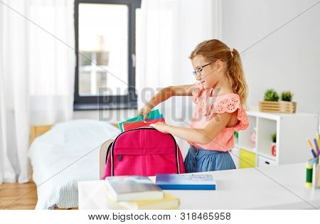 children, education and learning concept - happy student girl in glasses packing school bag at home
