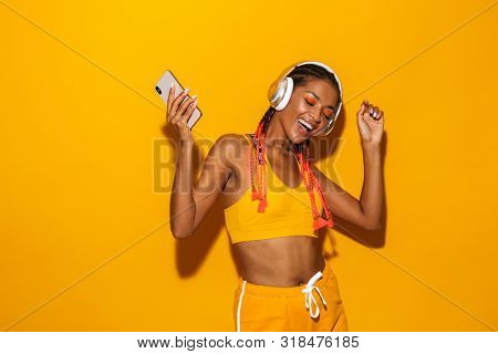 Image of beautiful african american woman singing while listening to music with smartphone and headphones isolated over yellow background