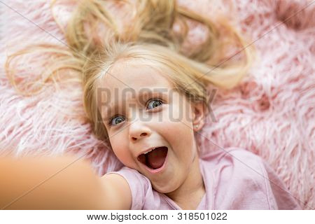 Adorable Little Girl With Long Blonde Hair Lying On Pink Fur Blanket And Making Selfie. Positive Emo