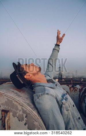 Urban Man With Hat Against The Background Of The City. Wearing White Shirt And Jeans. Old Neglected 