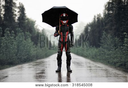 Motorcyclist In Full Gear And Helmet With Umbrella In The Rain. Motorcyclist In The Dark Woods.