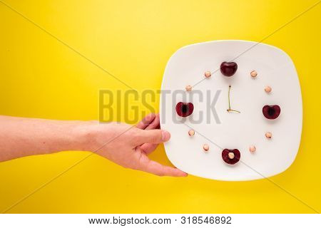 Clock Made Of Cherries On A Plate A Hand Is Reaching For On Yellow.