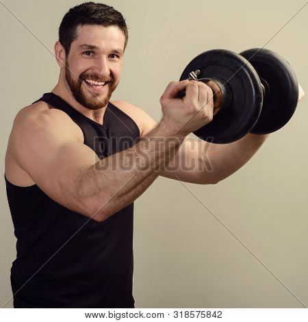 A Smiling Young Bearded Athlete In A Black T-shirt Raises A Heavy Dumbbell In Front Of Him.