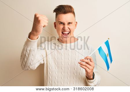 Young handsome man holding Argentinian Argentina flag over isolated white background annoyed and frustrated shouting with anger, crazy and yelling with raised hand, anger concept