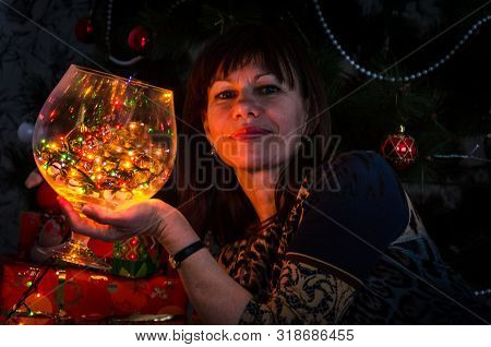 Close-up Portrait Of A Beautiful Adult Brunette Woman Near The Christmas Tree. Holds A Large Glass W