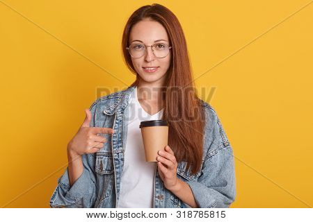 Image Of Young Dark Haired Female Pointing On Paper Coffee Cup Which Holds In Hand, Woman Dressed De