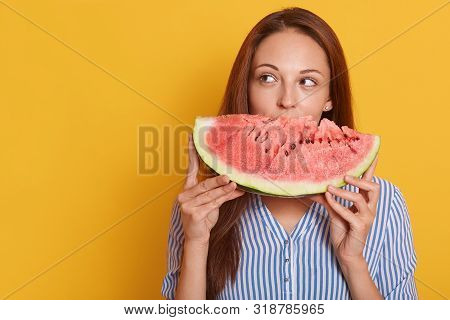 Indoor Shot Of Young Woman Biting Big Slice Of Watermelon While Looking Aside, Wearing Striped Shirt