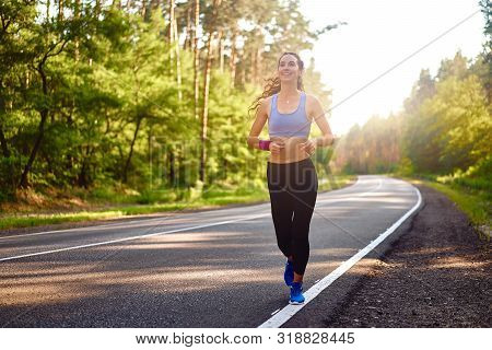 Beautiful Caucasian Young Girl Athlete Runs Sunny Summer Day On Asphalt Road In The Pine Forest. Wom